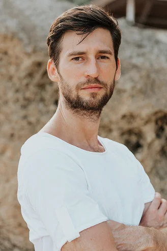 Een portret van een man met een baard en kort bruin haar, die met de armen over elkaar staat in een wit T-shirt met een natuurlijke, rotsachtige achtergrond