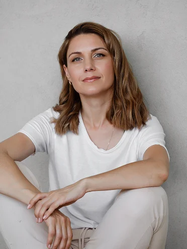 Een portret van een rustig kijkende vrouw met halfbruin haar, die ontspannen op de grond zit in een lichtroze T-shirt en bijpassende broek tegen een zachtroze muur